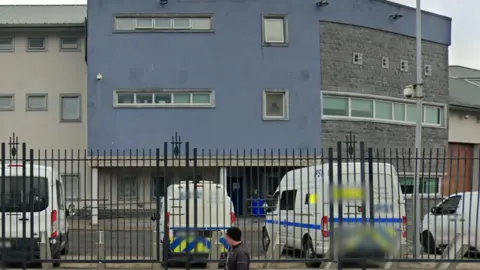 Google A white wall prison with three stories and blue front. Six vehicles are parked outside, including a police car. 