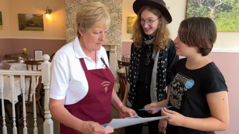 Helen is stood in the middle, looking down at a piece of paper she is holding, while Isaac and Imogen, stood next to her, talk about their project. They are stood in an old fashioned tea room.
