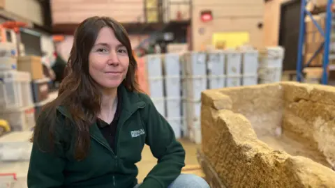 Emma Baugh/BBC Jessica Lowther crouching next to a stone Roman sarcophagas, which does not have a lid, and has some wear/damage on the top of one side.