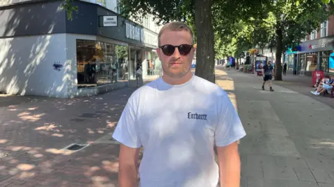 Phil Harrison/BBC A man wearing sunglasses and a white shirt. He is standing on a high street with shops and trees in the background.