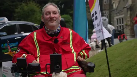 Steven Webb is sitting in an electric wheelchair. He is wearing a red Cornwall Air Ambulance outfit. Next to him is a flag flying in the wind which has a Cornwall flag on it. 