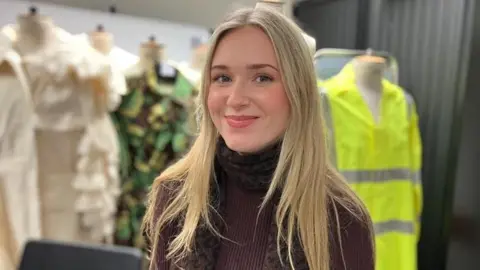 Alex Dunlop/BBC Hollie Ralph has long blonde hair and blue eyes and wears a brown polo neck jumper and a patterned scarf. Behind her are several mannequins displaying different outfits, including a cream-colored ruffled garment, a camouflage-patterned outfit with leaf-like details and bright yellow high‑visibility jacket.
