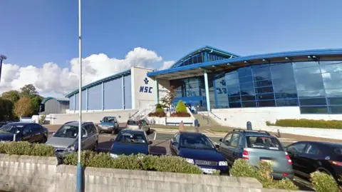 The exterior of the NSC, which is a large glass fronted building with a curved blue roof and a lettering that reads NSC a white wall next to the entrance. There are cars parked in front of the building.