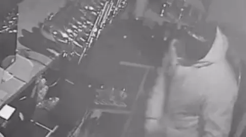 The Claydon Greyhound A still from grainy CCTV footage showing a masked intruder wearing a balaclav and a hooded coat while standing behind a pub bar.