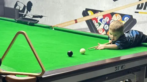 PA Media Jude, aged two, who has light brown hair and is wearing a blue t-shirt and black trousers, leans over the edge of a snooker table with a cue in his hands and aims at a white ball. A black ball is a few inches ahead. 