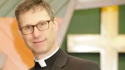 Diocese of Blackburn An image of Bishop Philip wearing a clerical collar and black jacket, he has short mousey brown hair and wears glasses as he smiles at the camera