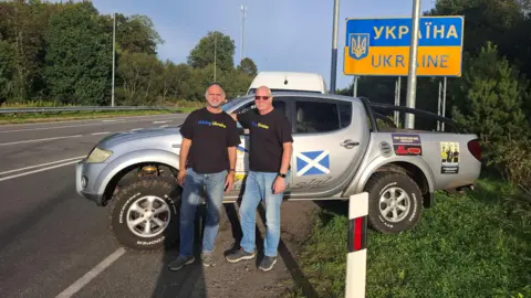 Steve Eccleshall Two men stood in front of a pick-up truck next to a sign marking the Ukrainian border