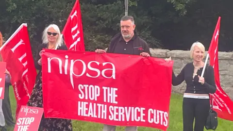 Two women and a man are pictured. The man has dark hair and a greying beard and is holding a red banner with white reading that says: "nipsa, Stop the health service cuts." On either side of him, is a woman. The woman on the left has blonde hair and sunglasses and is wearing a long dress. the woman on the right is wearing a dark top and glasses and has short, blonde hair. She is holding a red banner.