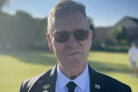 Adam Marchbank standing in front of a bowling green, wearing a jacket, shirt and tie, and sunglasses.