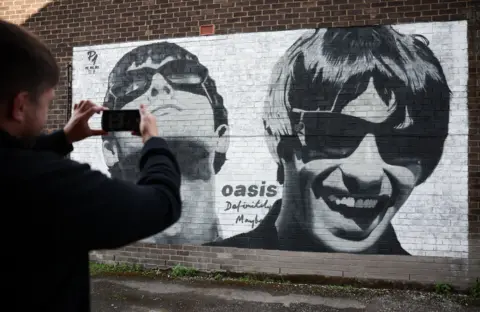 EPA Seorang penggemar memotret mural Liam dan Noel Gallagher dari band Inggris Oasis di dekat Sifters Records di Manchester, Inggris, 29 Agustus 2024. Mural tersebut dilukis oleh seniman jalanan Pic.One.Art pada bulan Juni 2024 untuk menandai ulang tahun ke-30 album perdana band tersebut, Definitely Maybe. 