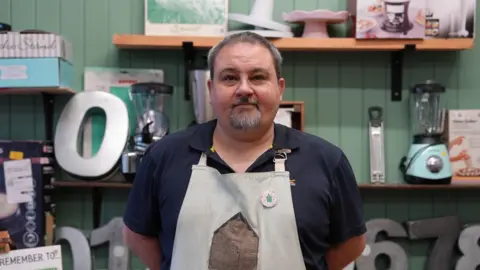 Dawid Wojowitz/BBC The manager of the store, Ben Mair, stands in front of the kitchen and baking section of the store. He's wearing a navy blue polo shirt and a pale blue apron with a brown shed on it. Ben has a greyish goatee and short hair. He's looking directly at the camera