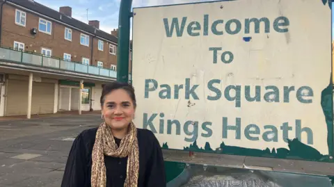 Rufia Ashraf with short dark hair wearing a black top with a brown and yellow scarf. She is standing in front of a sign that says "Welcome to Park Square, Kings Heath". A boarded-up shop is visible in the background.