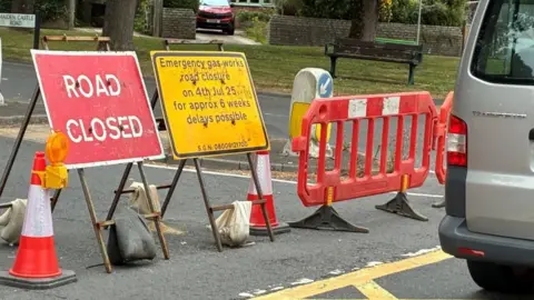 A red road closed sign and a yellow sign stating: "Emergency gas works road closure on 4th Jul 25 for approx 6 weeks, delays possible", can be seen at the end of Maiden Castle Road. There is also a red barrier and cones.