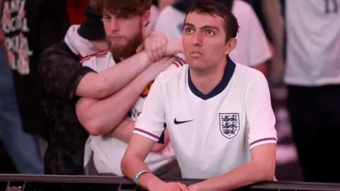 PA Media A fan wearing an England shirt leans on a barrier and stares up at a screen, while two other fans console each other