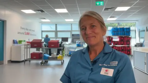 Image shows Vanessa smiling to the left of the photo. She is wearing a blue top which has a badge on it. Her hair is tied back. Behind here, are red and blue bags where the donated blood is packed before being taken to hospitals. 