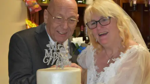 Steven and Julie Murfin cut the cake on their wedding day. He wears a dark grey suit, white shirt and tie, with a white rose pinned to his lapel. She wears a wedding dress and veil.