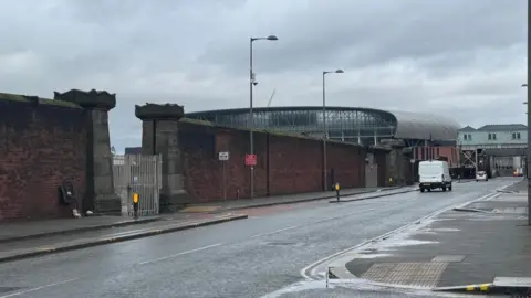 A view towards Everton Stadium on a road lined by a large wall on one side and former dockland buildings on the other