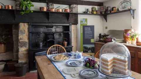 National Trust An old kitchen in the manor - large black iron stove at the back with copper kettles. Large pine cabinets to the side. Shelves above a beam above the stove with copper pans and vintage food tins. In the foreground, a traditional pine table with blue antique crockery and a fake display cake.