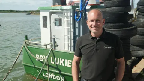 John Fairhall/BBC A man in a black polo shirt looks at the camera while standing in front of a green boat on the water.