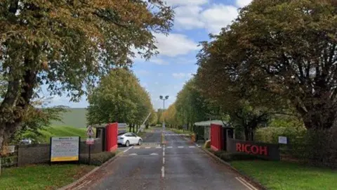 Google A long road stretches into the distance with green grass and trees on either side of it. There is a brick wall with bright red gates and a red and white barrier for vehicles beyond it. A white car is parked on the left. On the brick wall there is a red sign that reads "Ricoh"