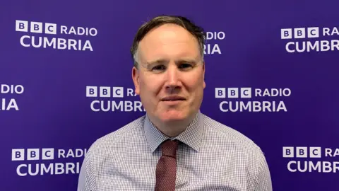 Owain Roberts has short grey/brown hair and is wearing a shirt and maroon tie. He is standing in front of a purple BBC Radio Cumbria branded backdrop.