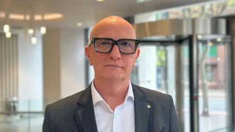 A bald man with black thick rimmed glasses. He is wearing a grey jacket and a white shirt.