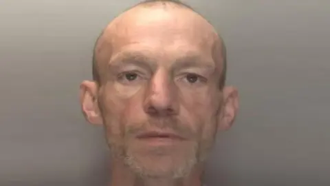 Baker appears in a police mugshot and has a shaved head and short ginger/blonde beard.
