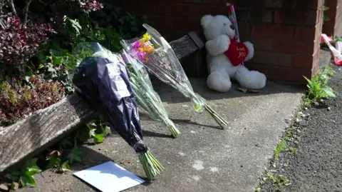 Three bunches of flowers, a white greetings card envelope and a white teddy bear carrying a red heart are lined up along a low fence at the edge of a pavement