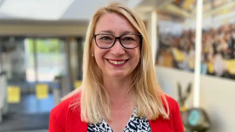 Jess Asato who is standing up while having her picture taken. She is looking directly at the camera and smiling and is wearing a white and black blouse with a red blazer over the top.