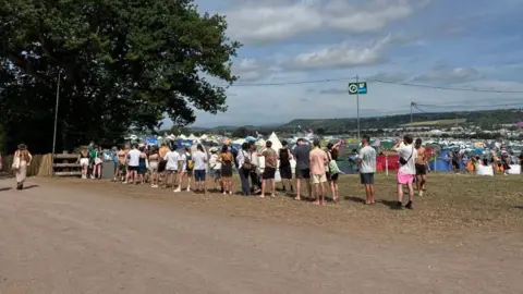 A queue of people waiting for their turn to fill up their water supply. There are tents in the background.