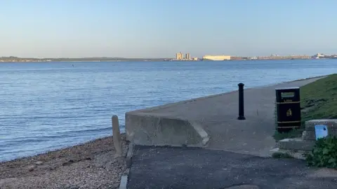 Road ending onto beach and sea beyond a promenade/path leads off the road with a black bin on the end water and docks beyond.