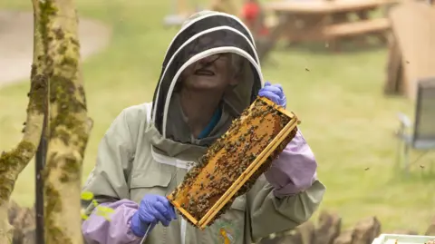 Gorman Photography A woman in a bee keeper suit is holding up bees while smiling at the camera.