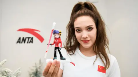 Freestyle skier Eileen Gu posing for photos in front of the Anta logo on a wall, holding a mini figurine of herself
