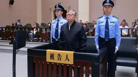 Yang Youlin appeared in court wearing a dark jacket standing between two uniformed police officers