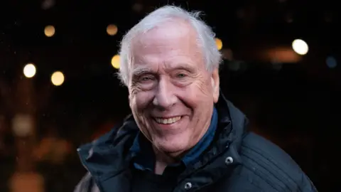 A close-up of Martin Tyler, wearing a dark blue shirt and black coat, smiling at the camera
