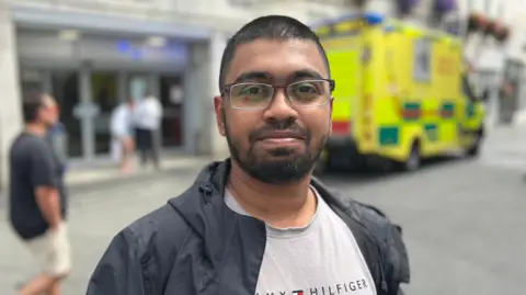 Sheikh Sakib - A brown skinned man with black hair, a black beard with glasses. He's wearing a black rain coat and a browny grey shirt. 