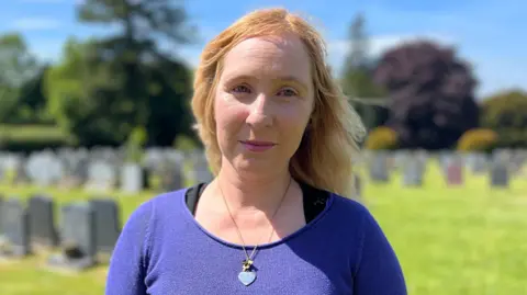 A woman with fair hair is looking into the camera, she is wearing a heart pendant necklace, and a purple jumper. She is standing in a cemetery with rows of gravestones in the distance behind her. It is a sunny summer's day. 