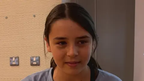 A girl called Rosie. She has black hair with a centre parting. It is tied up at the back. She has feckles on her face. She is wearing a blue shirt.