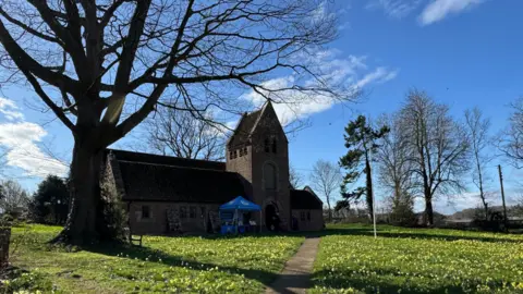 The image shows St Edward's Church in Kempley, Gloucestershire. A blue gazebo stands outside. The land surrounding the church is covered in grass and daffodils.