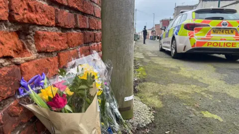 Photo of bouquet of flowers on the ground in alleyway by a brick wall. On the road is the back of a police car. The sky is grey.