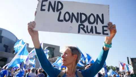a woman holds a sign saying no london rule as indy supporters hold saltires behind her