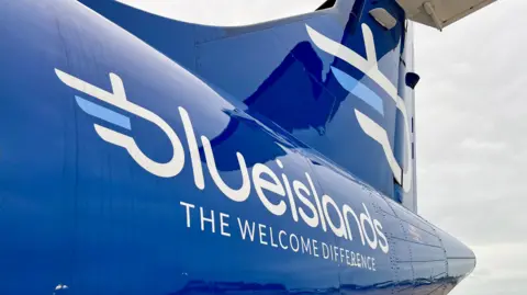 BBC A close up picture of the rear end of the outside of a Blue Islands plane. It is a royal navy blue colour with the white company logo prominent. 