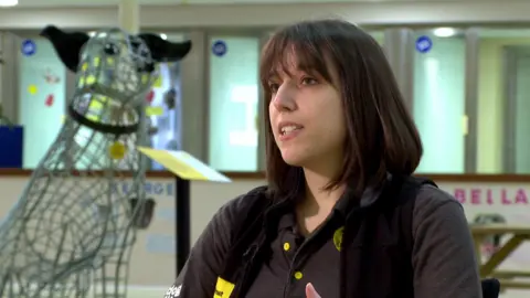 Emma Wakefield , who has dark brown hair and is wearing a Dogs Trust uniform in conversation. There is a large metal wire sculpture of a dog with black ears and a collar in the shape of the Trust's logo in the background.