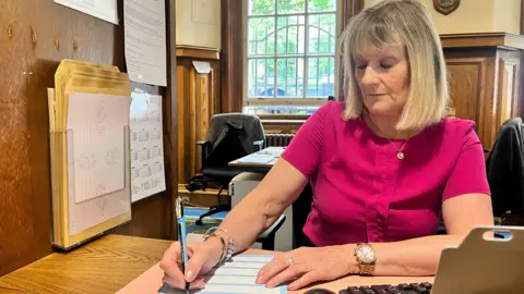 Jennifer is signing papers in her office. She has short blonde hair with a fringe and is wearing a pink blouse and a gold watch.