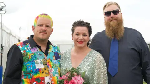 Eric Hobson On the left is a man wearing a black shirt and a colourful patch waistcoat with lots of eccentric designs. His short hair is dyed in lots of neon colours. He is standing next to a woman wearing a pale green sequin dress holding a bouquet of flowers, she has black hair and red lipstick and is smiling. On the right is a man with a large beard and sunglasses wearing all black shirt with a navy tie.