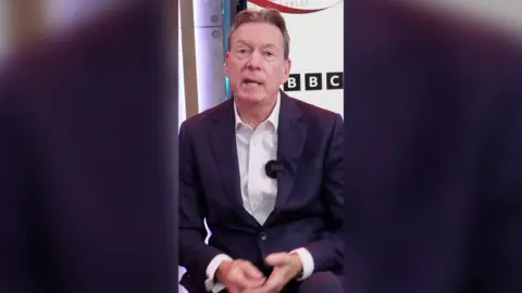 Frank Gardner is sat down and wearing a navy suit with a white shirt. Behind him is a screen with the BBC logo on.