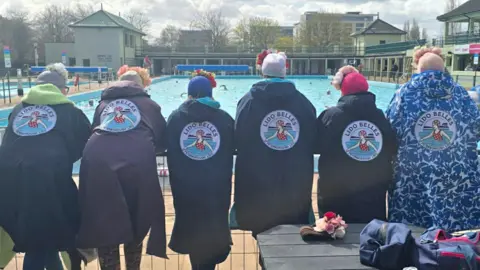 Submitted A line of six women look out at the lido. They have their backs to the camera. They are all wearing woolly hats and waterproof jackets bearing the circular Lido Belles logo.