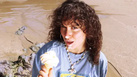 Maximilliaan van Es A young-woman with brown curly hair and a fringe, holding an ice cream in a cone with a confused expression on her face. She has ice cream on her nose, lips and chin. She is wearing a light purple sweatshirt, with a flower necklace on made of shells. She is wearing large hoop earrings and is stood on a beach with a rock to her left and wet sand behind her.