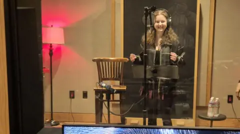 Erin Rose stood in a recording booth. She has long dark curly hair and is wearing black headphones, a black top, black jacket and burgundy skirt. She is stood behind a microphone and is smiling.