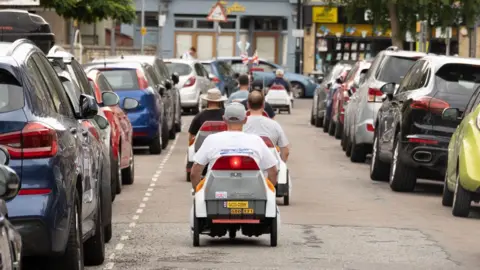 ARU About six Sinclair C5 vehicles are seen from behind in a row as they travel down a residential street. There are cars parked on both sides of the street and some shops in the background